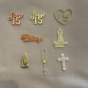 May include: Assortment of gold and white cake toppers with various designs, including "Sweet 15", "Feliz Bautizo", "Nuestra Boda", a heart with "15" inside, a praying hands, a rosary, and a cross.