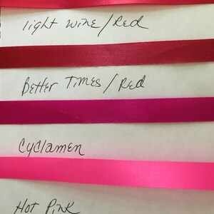 May include: Four horizontal satin ribbons in shades of red and pink arranged on a white surface. The ribbons are labeled with handwritten text: light wine/red, Better Times/red, Cyclamen, and Hot Pink, corresponding to each color strip.