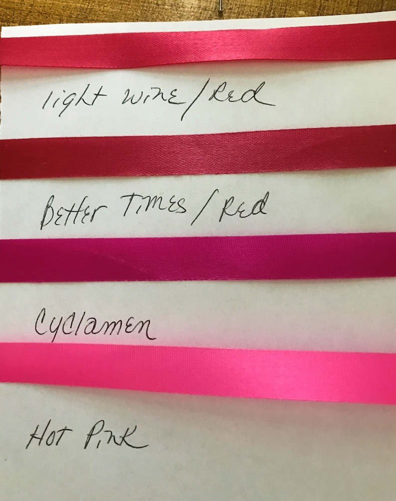 May include: Four horizontal satin ribbons in shades of red and pink arranged on a white surface. The ribbons are labeled with handwritten text: light wine/red, Better Times/red, Cyclamen, and Hot Pink, corresponding to each color strip.