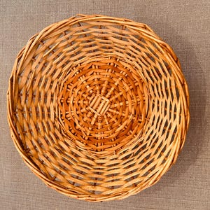 Puede incluir: Una cesta redonda y poco profunda construida con mimbre tejido de color marr&oacute;n claro. El dise&ntilde;o presenta una base circular con un patr&oacute;n radiante de radios y anillos tejidos conc&eacute;ntricos que se extienden hacia afuera formando los lados curvos.
