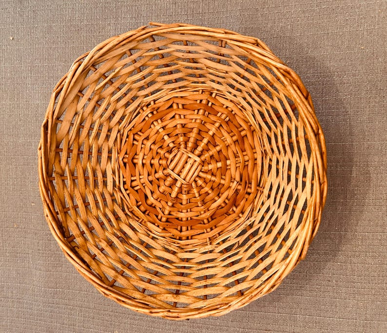 Puede incluir: Una cesta redonda y poco profunda construida con mimbre tejido de color marr&oacute;n claro. El dise&ntilde;o presenta una base circular con un patr&oacute;n radiante de radios y anillos tejidos conc&eacute;ntricos que se extienden hacia afuera formando los lados curvos.