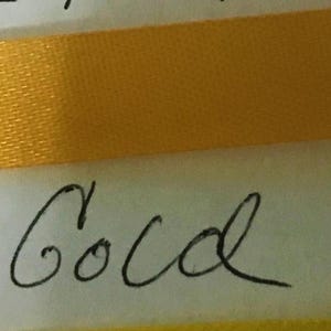 May include: A horizontal strip of smooth, shiny gold ribbon positioned across a white surface. Below the ribbon, the words "old Gold" are written in black cursive script. The ribbon has a consistent texture and a vibrant, reflective yellow-gold color.
