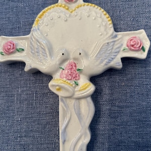 May include: White ceramic cross with a heart, two doves, and pink flowers. The doves are holding a bouquet of pink flowers and two bells.