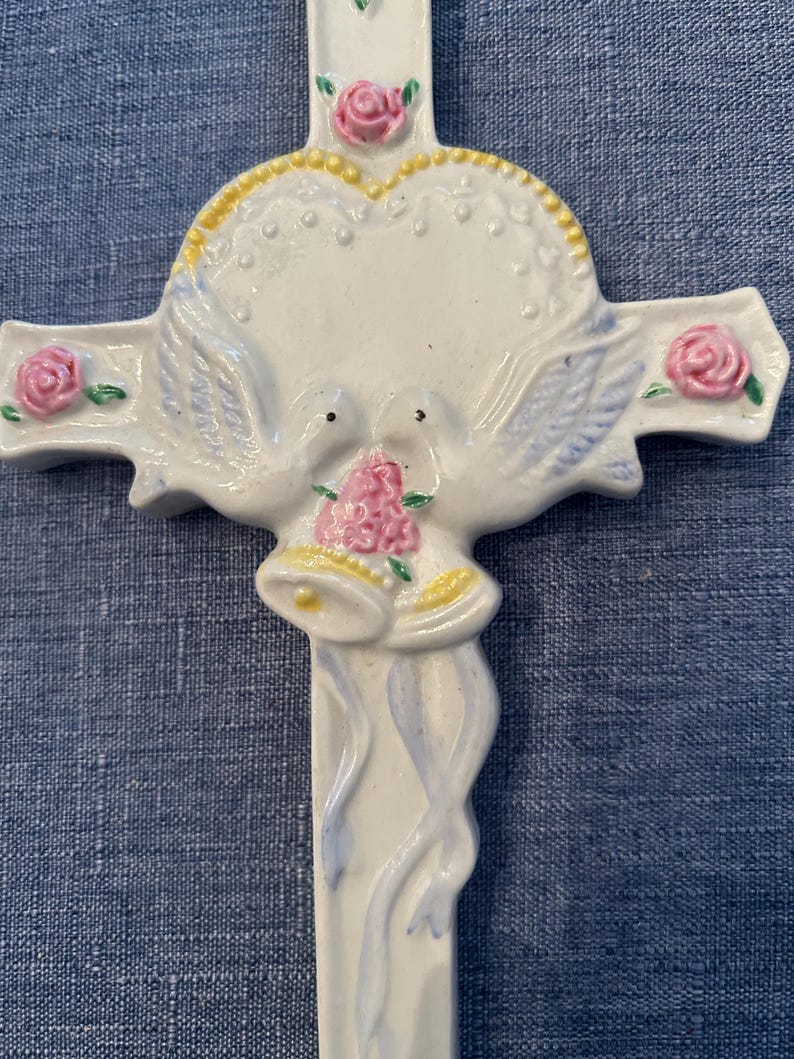 May include: White ceramic cross with a heart, two doves, and pink flowers. The doves are holding a bouquet of pink flowers and two bells.