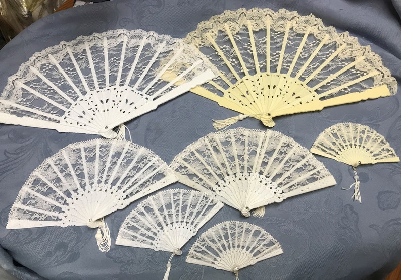 May include: A collection of eight white and ivory lace fans. The fans are made of delicate lace and have a variety of designs. Some of the fans have tassels.
