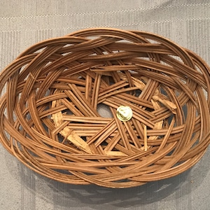 May include: Oval-shaped woven basket made of light brown wicker. The basket has a central opening and a gold-colored pin in the center. The basket's design features interwoven strips of wicker, creating a textured surface. The basket is likely used for storage or display.