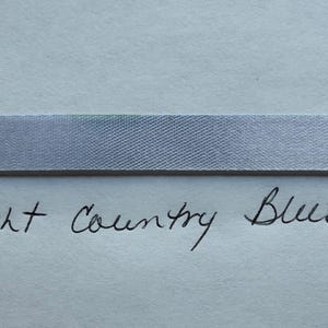 May include: A horizontal strip of light blue ribbon with a smooth, woven texture. Below the ribbon, the words light Country Blue are handwritten in black ink on a plain, light-colored surface, clearly identifying the color of the ribbon.