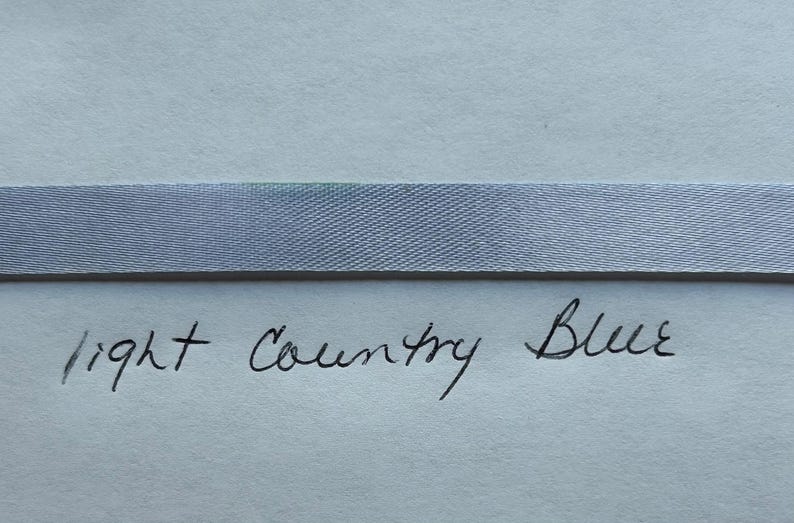 May include: A horizontal strip of light blue ribbon with a smooth, woven texture. Below the ribbon, the words light Country Blue are handwritten in black ink on a plain, light-colored surface, clearly identifying the color of the ribbon.