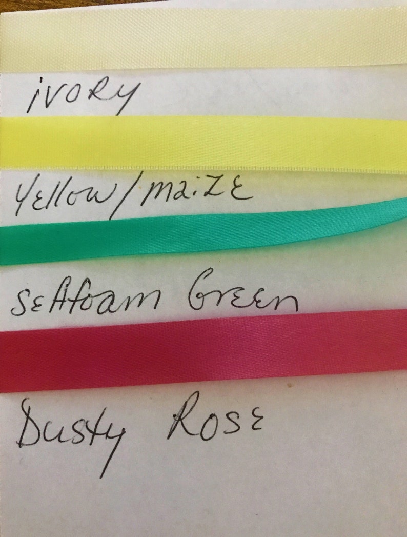 May include: Four horizontal strips of smooth satin ribbon in ivory, yellow, seafoam green, and dusty rose colors. Each ribbon is labeled with handwritten text below it, reading ivory, yellow/maize, seafoam green, and dusty rose respectively.