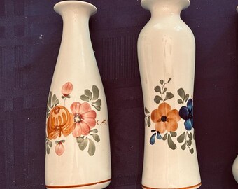 May include: Two off-white ceramic vases with narrow necks and flared rims. Each vase features hand-painted floral motifs in shades of orange, pink, blue, and green foliage. The bases are accented with a thin orange horizontal line around the bottom.