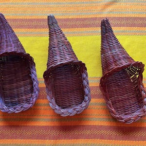 May include: Three dark brown woven wicker cornucopia baskets with a tapered, cone-like shape and a braided rim detail. Each basket features a wide, open front and a narrow, pointed back, constructed from thin, flexible reed material.