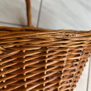 May include: Close-up of a woven wicker basket with a handle. The basket is made of light brown wicker and has a textured surface. The weave pattern is visible, and the handle is curved.