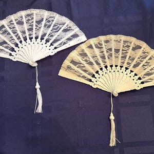 May include: Two white lace hand fans with tassels. The fan on the left is white and the fan on the right is a cream color.