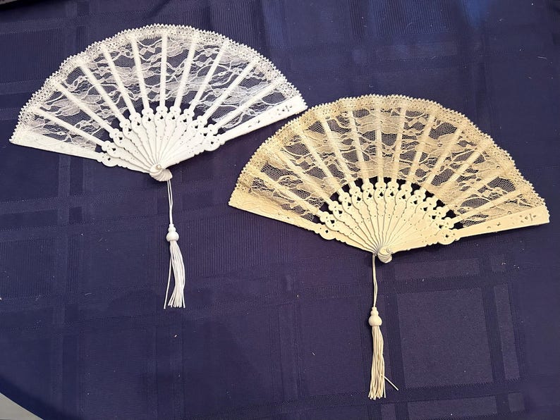 May include: Two white lace hand fans with tassels. The fan on the left is white and the fan on the right is a cream color.