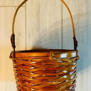 May include: A woven bamboo basket with a curved handle. The basket is a warm brown color with a glossy finish. The handle is attached with dark brown twine. The basket is designed with a rounded bottom and a flared top.