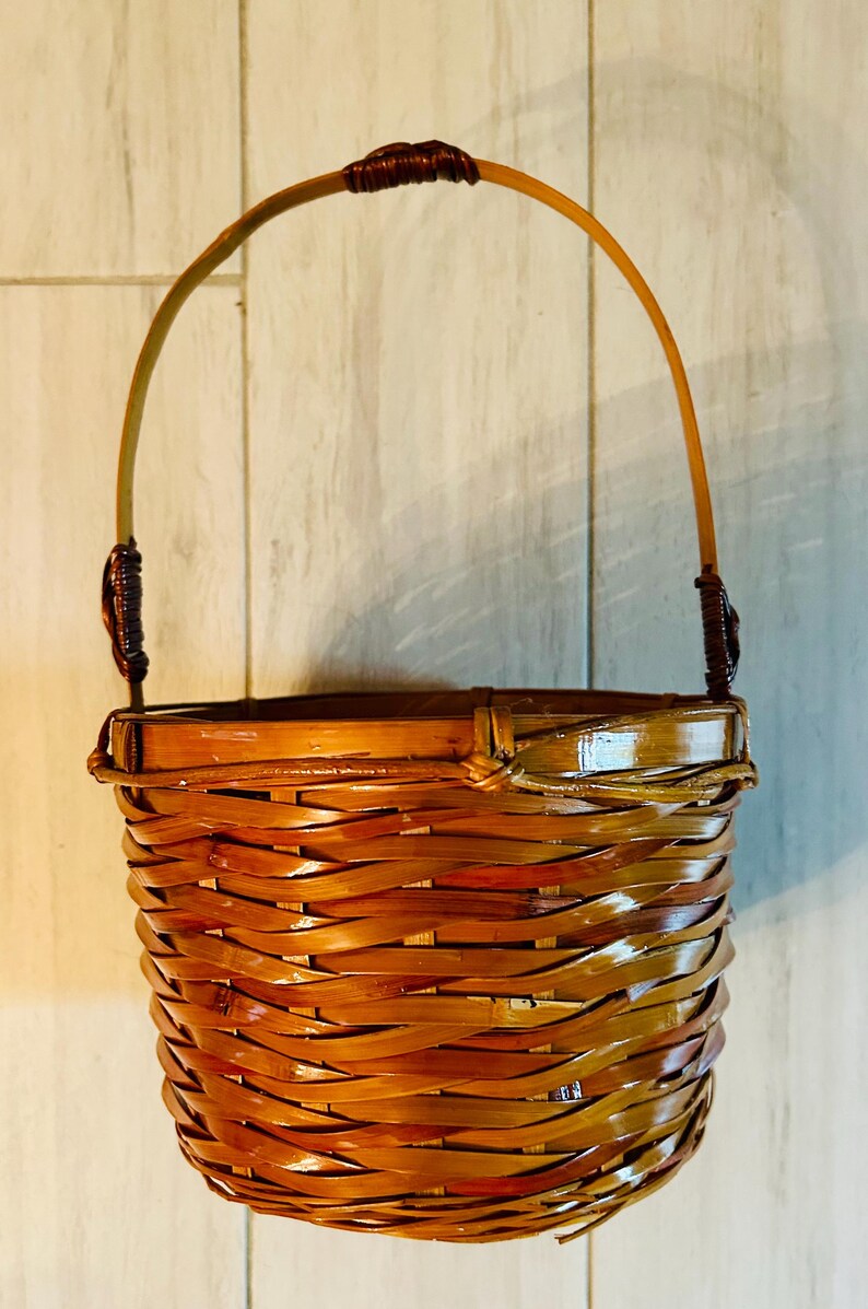 May include: A woven bamboo basket with a curved handle. The basket is a warm brown color with a glossy finish. The handle is attached with dark brown twine. The basket is designed with a rounded bottom and a flared top.