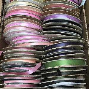 May include: A collection of numerous spools of satin ribbon in a wide variety of colors, including shades of pink, blue, green, yellow, purple, and black, all neatly stacked and organized within a rectangular cardboard storage box.