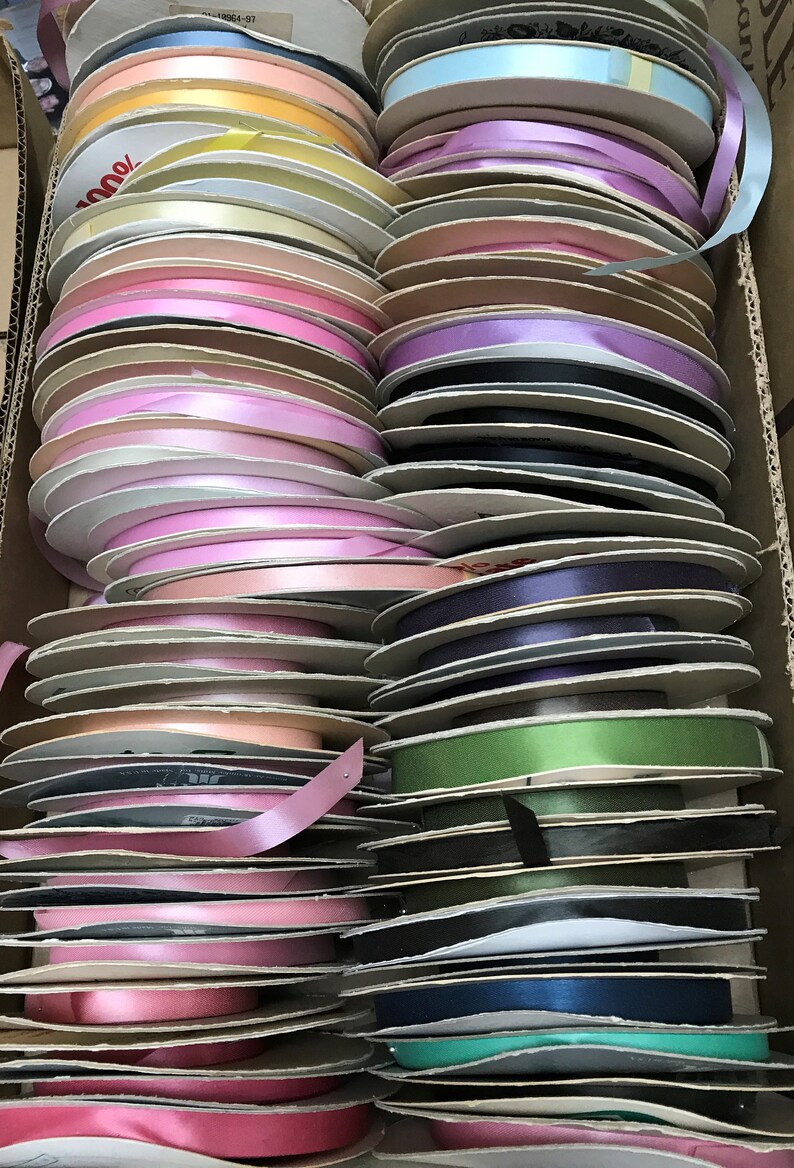 May include: A collection of numerous spools of satin ribbon in a wide variety of colors, including shades of pink, blue, green, yellow, purple, and black, all neatly stacked and organized within a rectangular cardboard storage box.
