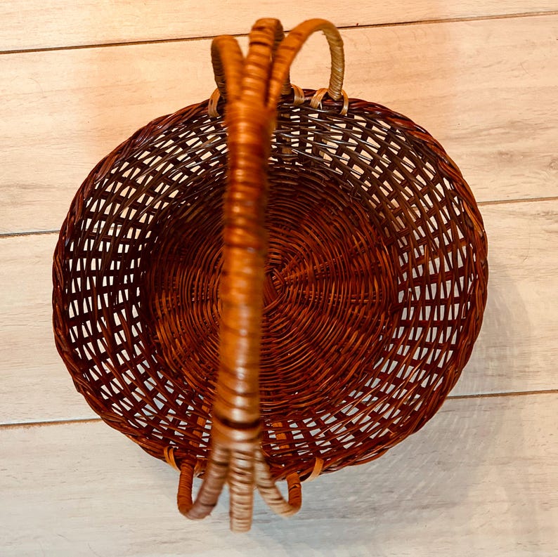 May include: A round, woven basket with a sturdy handle. The basket is made of brown wicker and has a circular design. The handle is attached to the basket with woven supports. The basket is empty.