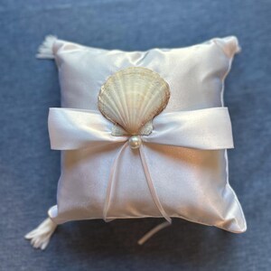 May include: A square white satin ring pillow featuring a central white ribbon tied in a bow. A natural tan seashell is attached to the center of the bow, accented by a small white pearl bead. The pillow has small white tassels at the corners.