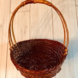 May include: A woven brown basket with a rounded base and a tall, arched handle. The basket is made of tightly woven, dark brown wicker, with a lighter-colored handle. The basket's interior is visible, showing the woven pattern.