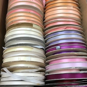 May include: A collection of satin ribbon spools arranged in two vertical columns within a cardboard box. The spools feature a variety of colors including shades of pink, peach, orange, yellow, white, lavender, purple, and magenta.