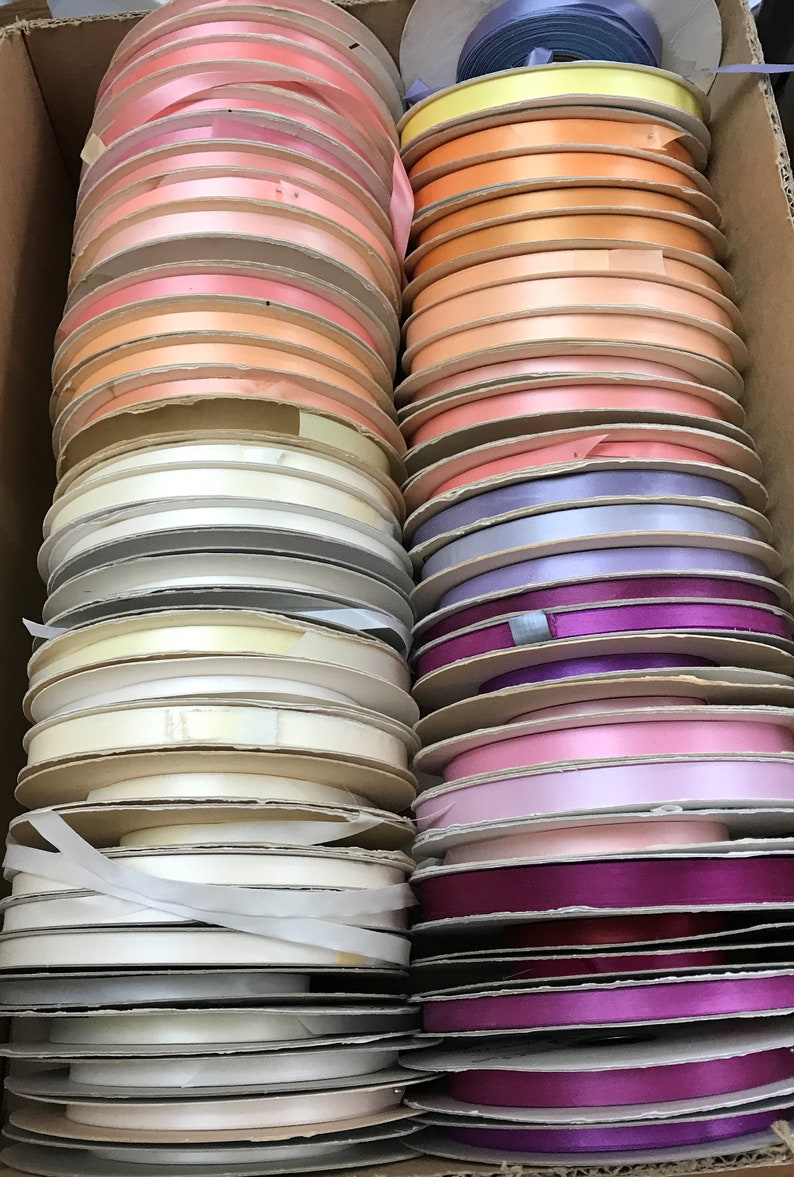 May include: A collection of satin ribbon spools arranged in two vertical columns within a cardboard box. The spools feature a variety of colors including shades of pink, peach, orange, yellow, white, lavender, purple, and magenta.
