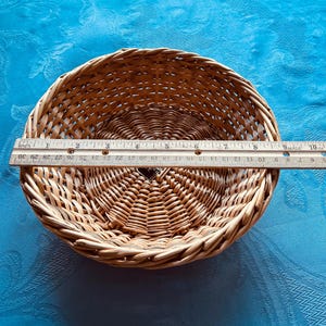 Puede incluir: Una cesta redonda de mimbre trenzado de color marr&oacute;n claro con base circular y borde trenzado. Una regla de madera est&aacute; colocada sobre el centro de la cesta, mostrando un di&aacute;metro de aproximadamente 24 cm. La cesta tiene una peque&ntilde;a etiqueta oscura.