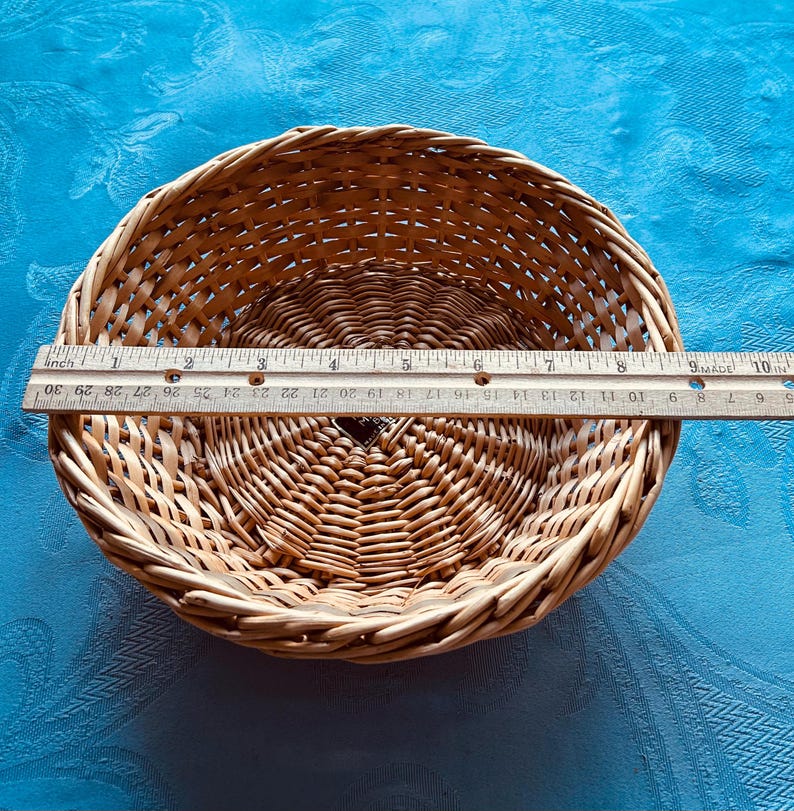 Puede incluir: Una cesta redonda de mimbre trenzado de color marr&oacute;n claro con base circular y borde trenzado. Una regla de madera est&aacute; colocada sobre el centro de la cesta, mostrando un di&aacute;metro de aproximadamente 24 cm. La cesta tiene una peque&ntilde;a etiqueta oscura.