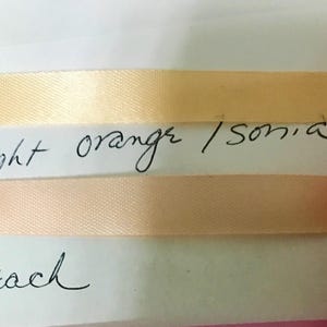 May include: Two strips of smooth, satin-like ribbon attached to a white card. The top ribbon is a light orange color with the handwritten text light orange / sonia below it. The bottom ribbon is a soft peach color with the word peach written below.