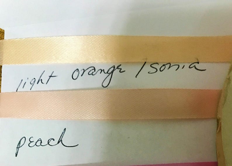 May include: Two strips of smooth, satin-like ribbon attached to a white card. The top ribbon is a light orange color with the handwritten text light orange / sonia below it. The bottom ribbon is a soft peach color with the word peach written below.