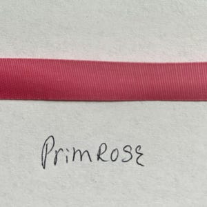 May include: A horizontal strip of solid dark pink ribbon with a smooth, woven texture. Below the ribbon, the word Primrose is handwritten in black ink on a plain, light-colored surface. The ribbon has a consistent width and matte finish.