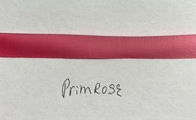 May include: A horizontal strip of solid dark pink ribbon with a smooth, woven texture. Below the ribbon, the word Primrose is handwritten in black ink on a plain, light-colored surface. The ribbon has a consistent width and matte finish.