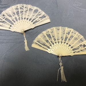 May include: Two white lace fans with ivory handles and white tassels. The fans are closed and have a delicate floral pattern.