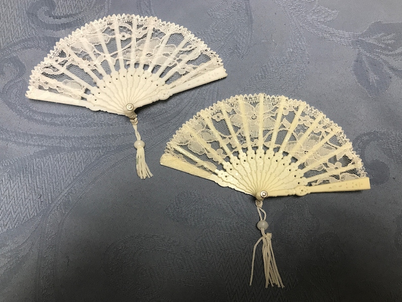 May include: Two white lace fans with ivory handles and white tassels. The fans are closed and have a delicate floral pattern.