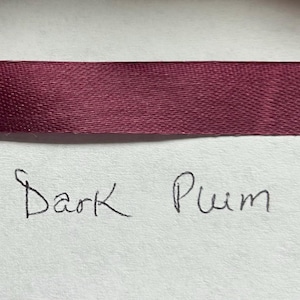 May include: A horizontal strip of dark plum colored satin ribbon with a smooth, reflective texture. Below the ribbon, the words Dark Plum are handwritten in black ink on a plain, light-colored surface, clearly labeling the color of the ribbon.
