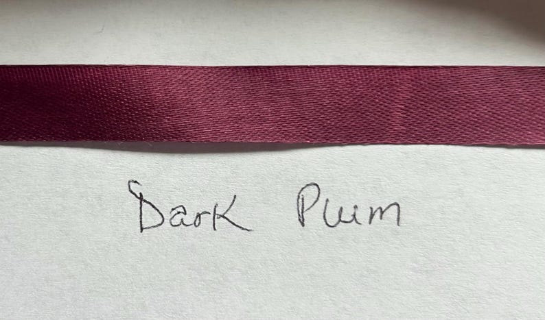 May include: A horizontal strip of dark plum colored satin ribbon with a smooth, reflective texture. Below the ribbon, the words Dark Plum are handwritten in black ink on a plain, light-colored surface, clearly labeling the color of the ribbon.