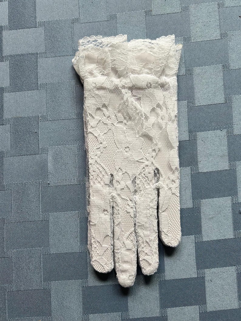 May include: A pair of white lace gloves with a delicate floral pattern. The gloves have a ruffled edge at the wrist.