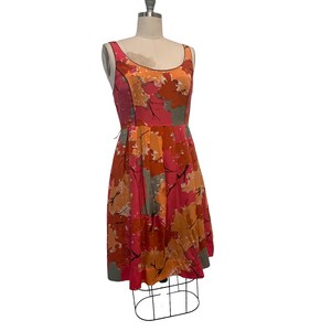 K&ouml;nnte beinhalten: Ein &auml;rmelloses Kleid mit einem abstrakten Bl&auml;tterdruck in Rosa, Orange und Grau. Das Kleid hat einen figurbetonten Oberteil und einen vollen Rock.