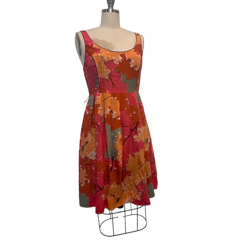 K&ouml;nnte beinhalten: Ein &auml;rmelloses Kleid mit einem abstrakten Bl&auml;tterdruck in Rosa, Orange und Grau. Das Kleid hat einen figurbetonten Oberteil und einen vollen Rock.