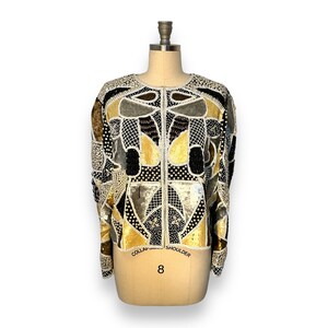 May include: A black, gold, and white sequined jacket with a geometric pattern. The jacket has a round neckline and long sleeves.