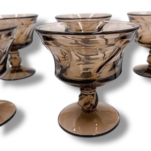 May include: Set of six vintage brown glass goblets. Each goblet features a wide, fluted bowl with a decorative swirl pattern and a short, twisted stem. The base is round and the glass has a warm, amber hue. Ideal for serving beverages.