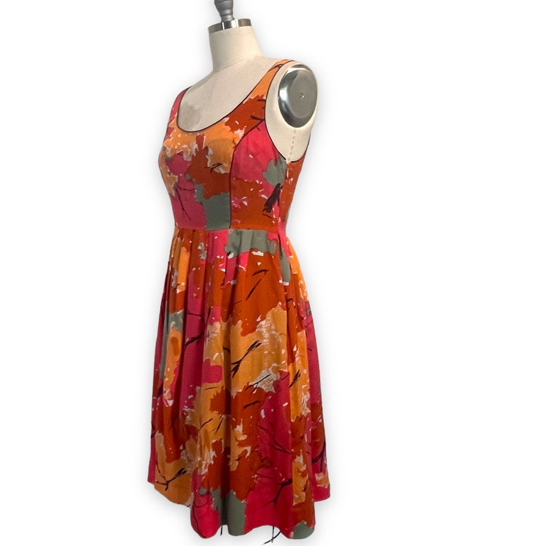 K&ouml;nnte beinhalten: Ein &auml;rmelloses Kleid mit floralem Druck in Rosa, Orange und Grau, mit einem runden Ausschnitt und einem vollen Rock.