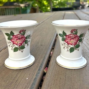 May include: Two white porcelain bud vases with a gold rim and base. Each vase features a large pink and red rose with green leaves. The vases are on a wooden surface.
