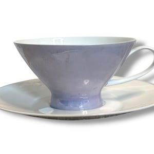 Pode incluir: Um conjunto de chávena e pires lavanda. A chávena tem uma forma larga e aberta com interior branco e uma pega curva. O pires é branco com uma borda ligeiramente levantada. O conjunto é projetado para serviço de chá.