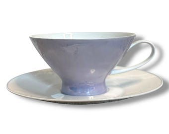 May include: A lavender teacup and saucer set. The teacup has a wide, flared shape with a white interior and a curved handle. The saucer is white with a slightly raised edge. The set is designed for tea service.