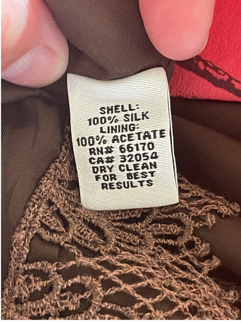 K&ouml;nnte beinhalten: Ein wei&szlig;es Etikett mit schwarzem Text, das die Stoffzusammensetzung eines Kleidungsst&uuml;cks beschreibt. Das Etikett lautet "SHELL: 100% SILK LINING: 100% ACETATE RN# 66170 CA# 32054 DRY CLEAN FOR BEST RESULTS".