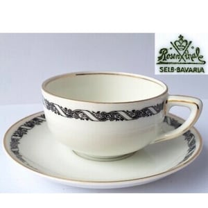 Puede incluir: Una taza de té y un platillo blancos con un diseño floral negro y dorado. El platillo tiene un borde dorado. La taza de té tiene un borde dorado y un diseño floral negro alrededor de la parte superior. La taza de té tiene un asa. El platillo tiene un diseño floral negro alrededor del borde. La taza de té y el platillo están hechos por Rosenthale Selb-Bavaria.