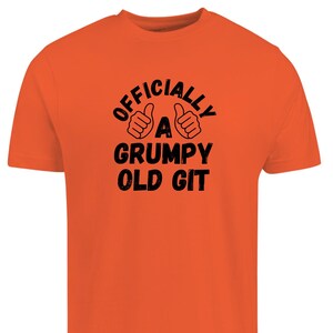 Può includere: T-shirt arancione con la scritta nera "OFFICIALLY A GRUMPY OLD GIT" e illustrazioni di pollici in su. La maglietta è a girocollo.