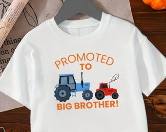 May include: White t-shirt with the text "PROMOTED TO BIG BROTHER!" in orange. The shirt features illustrations of a blue tractor and a red tractor with smoke. The shirt is hanging on a gray hanger.